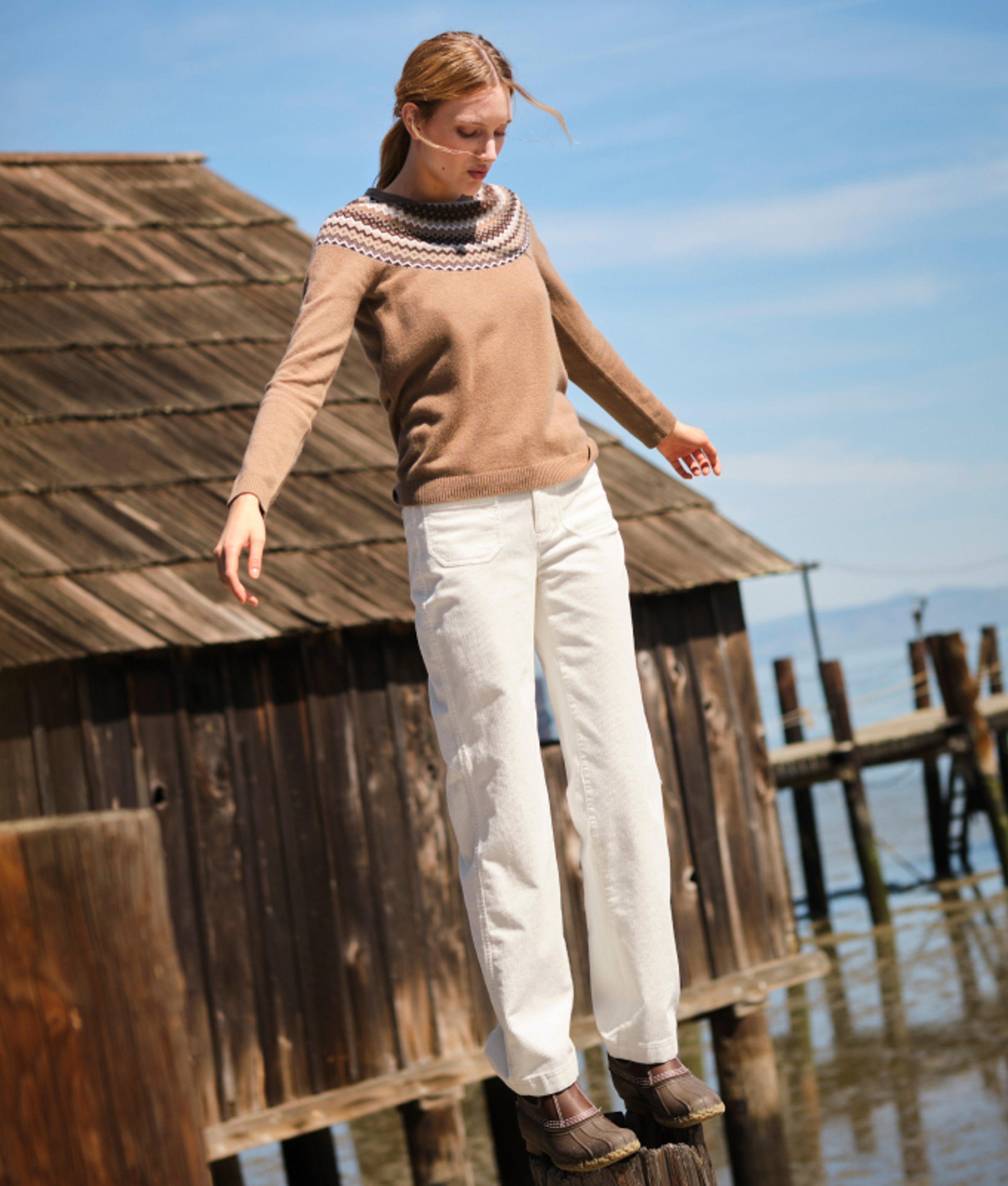 Woman balancing on a wooden post by a weathered waterside building.