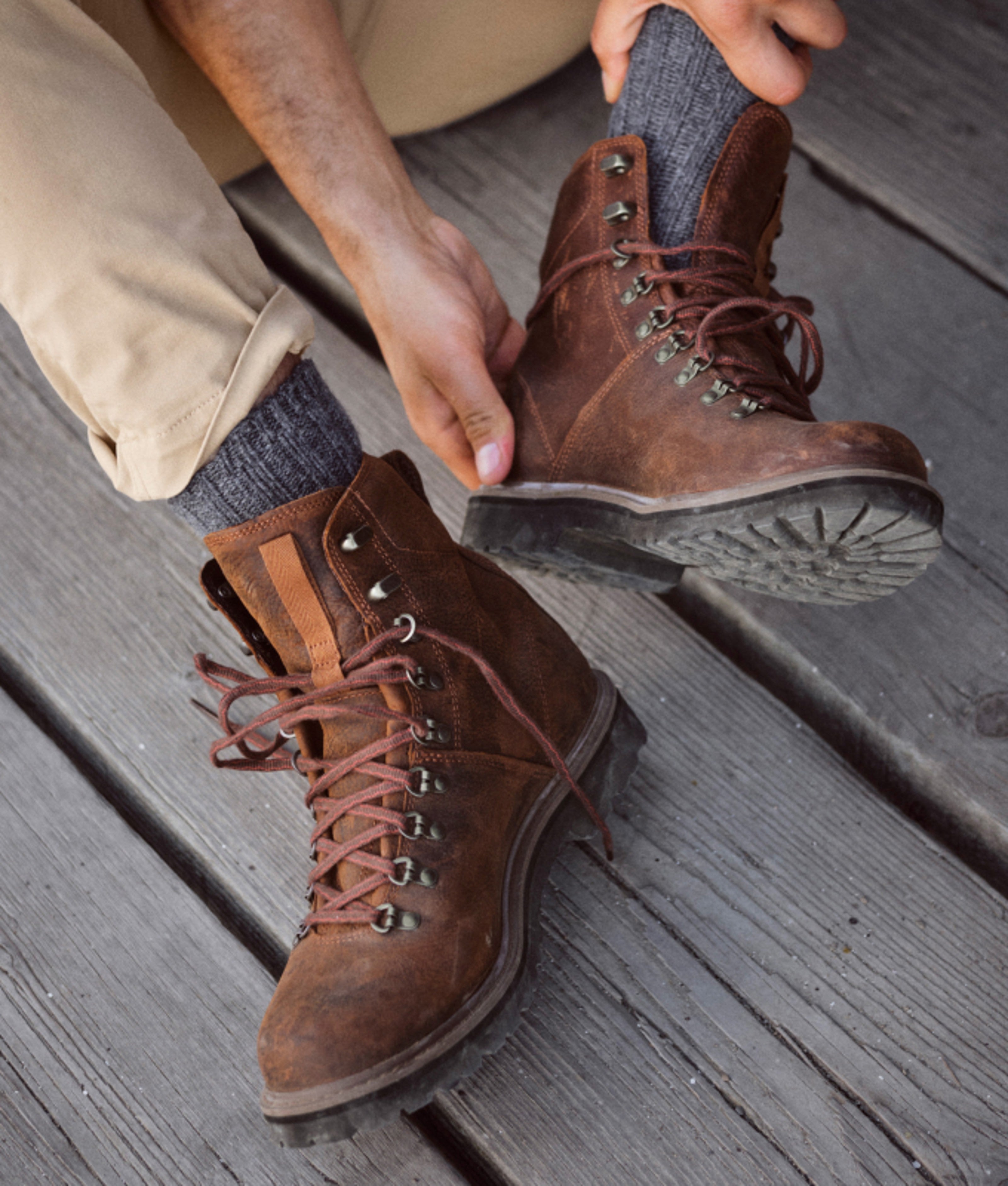 Hands lacing brown leather hiking boots on a wooden deck.